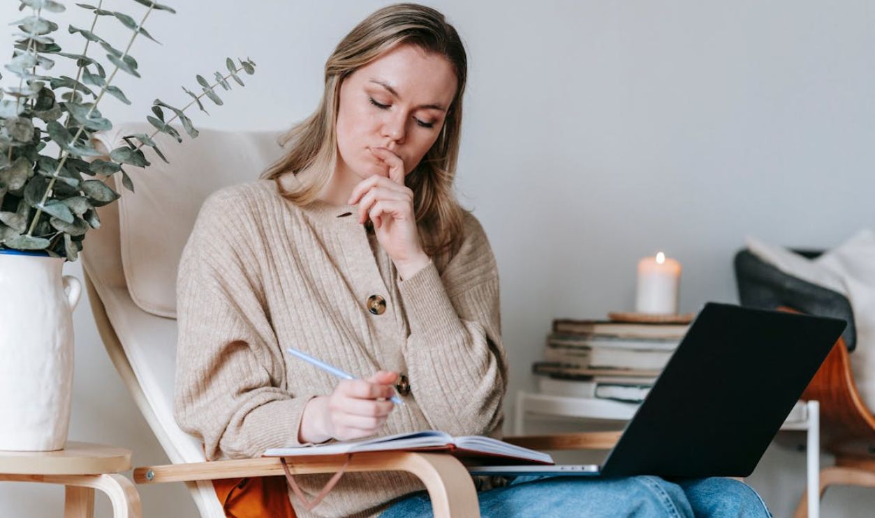 Thoughtful woman taking notes in planner while working with laptop