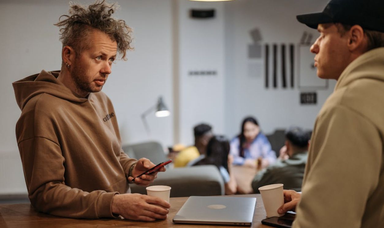 Two men in a casual office setting talking over coffee with laptops