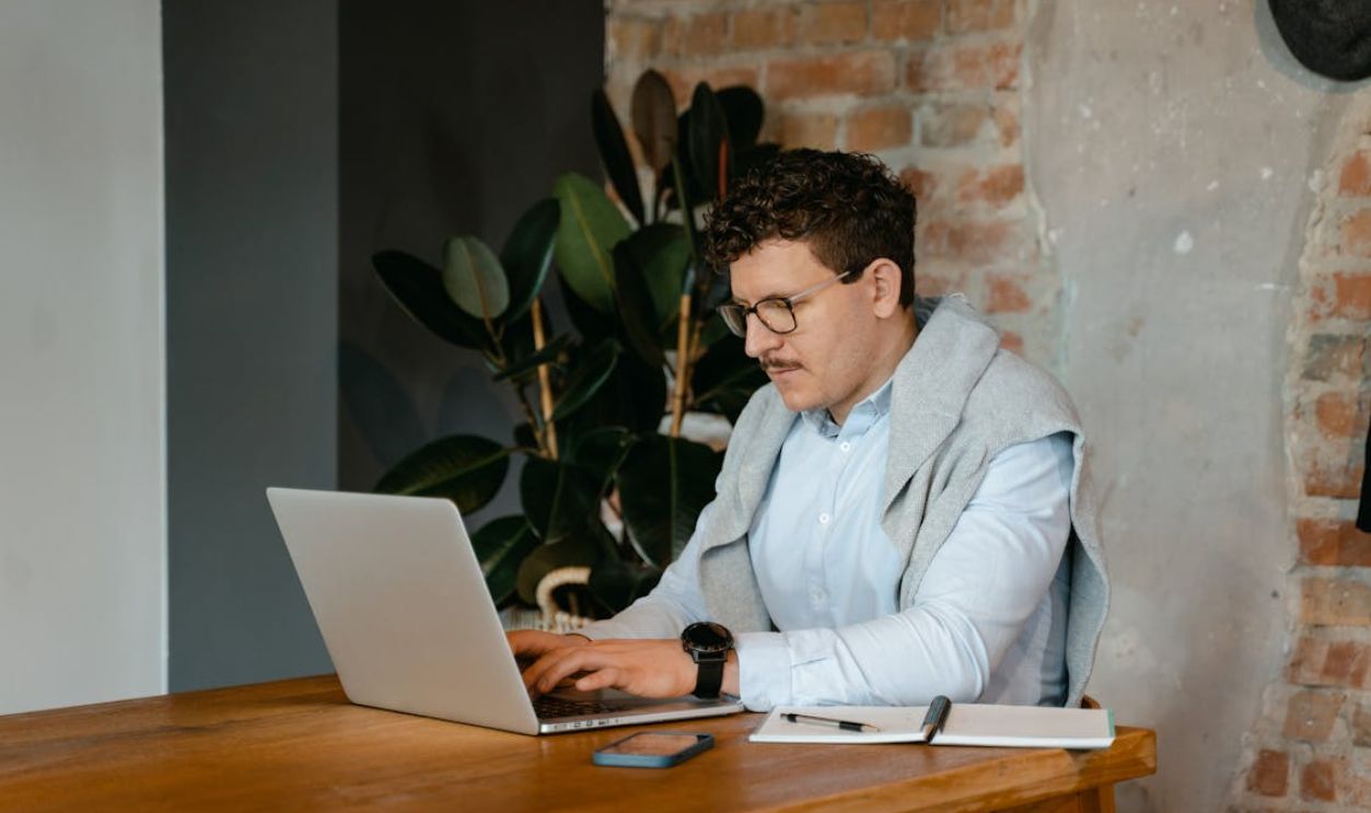 Adult man using a laptop at a wooden table in a cozy workspace with exposed brick walls