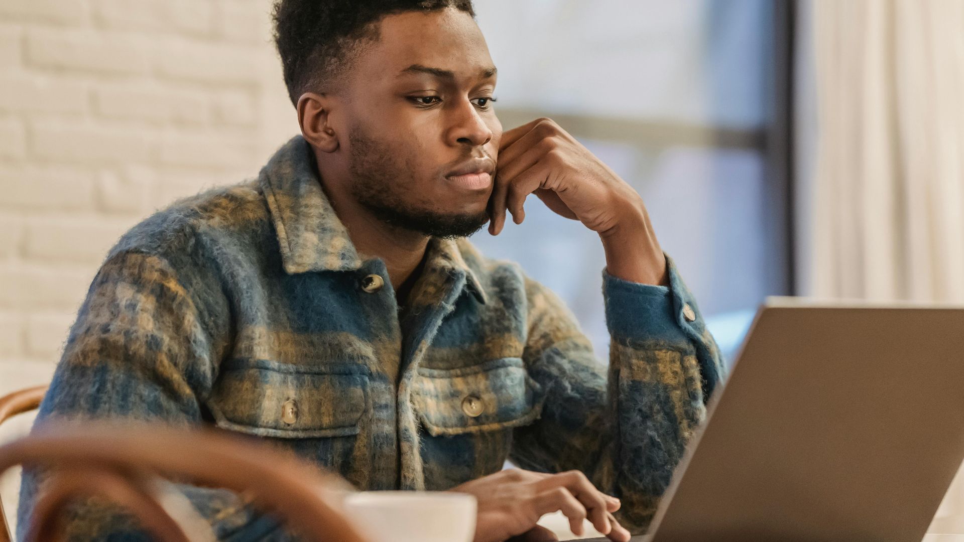 Concentrated bearded African American male freelancer in casual clothes sitting at table with laptop and leaning on hand while working on remote project in home workspace