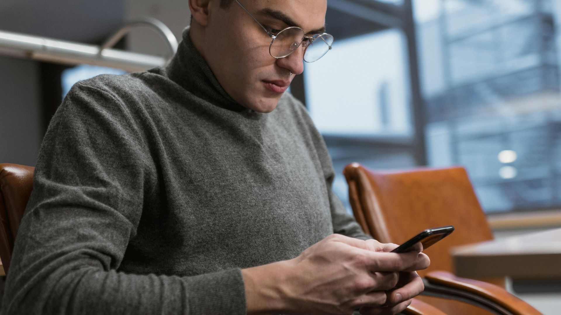 A young man in a grey sweater using a smartphone at an office desk.