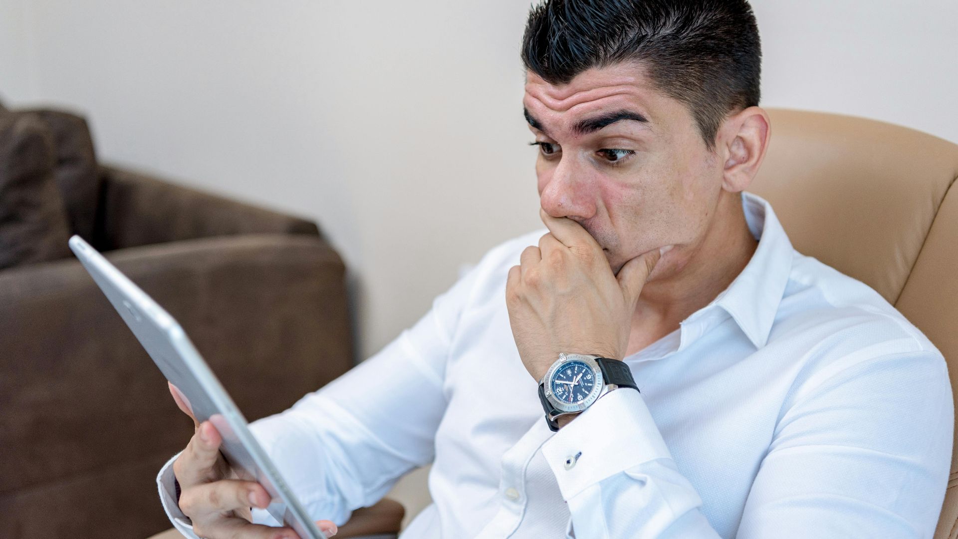 A man in a white shirt looks surprised while reading content on a tablet.