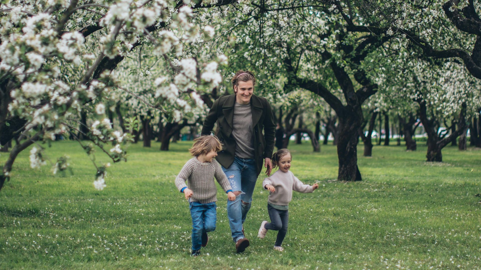 Father and children running joyfully in a lush, blossoming spring orchard.