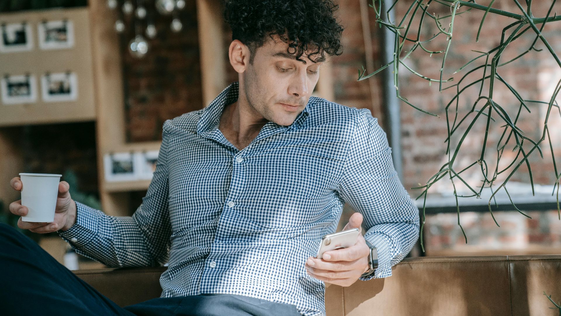 Man in stylish attire enjoying coffee while checking phone in a trendy cafe setting.