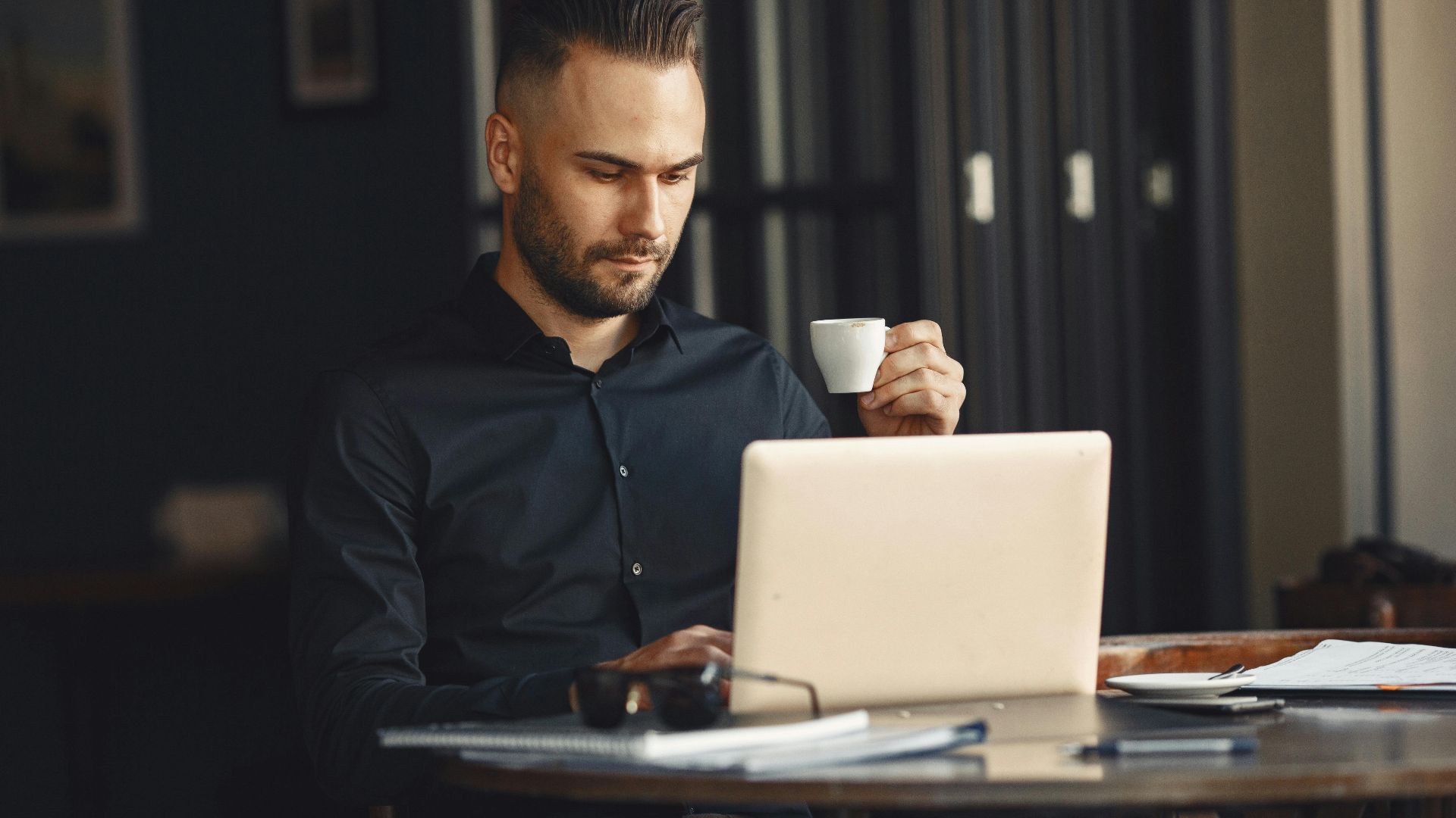 Adult man in a black shirt focused on his laptop, holding a coffee cup while working remotely indoors.
