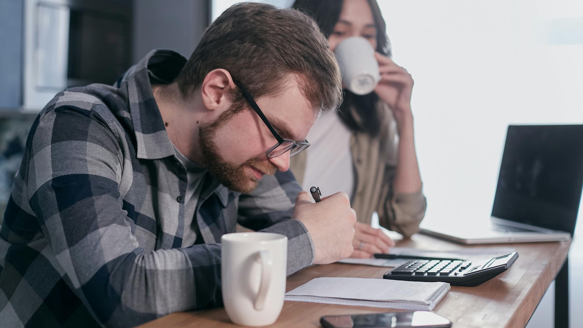 A couple sits at a kitchen table, reviewing finances with a laptop and calculator.