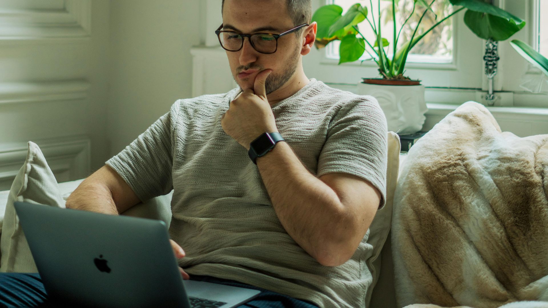 A man wearing eyeglasses using a laptop in a cozy home setting with natural light.