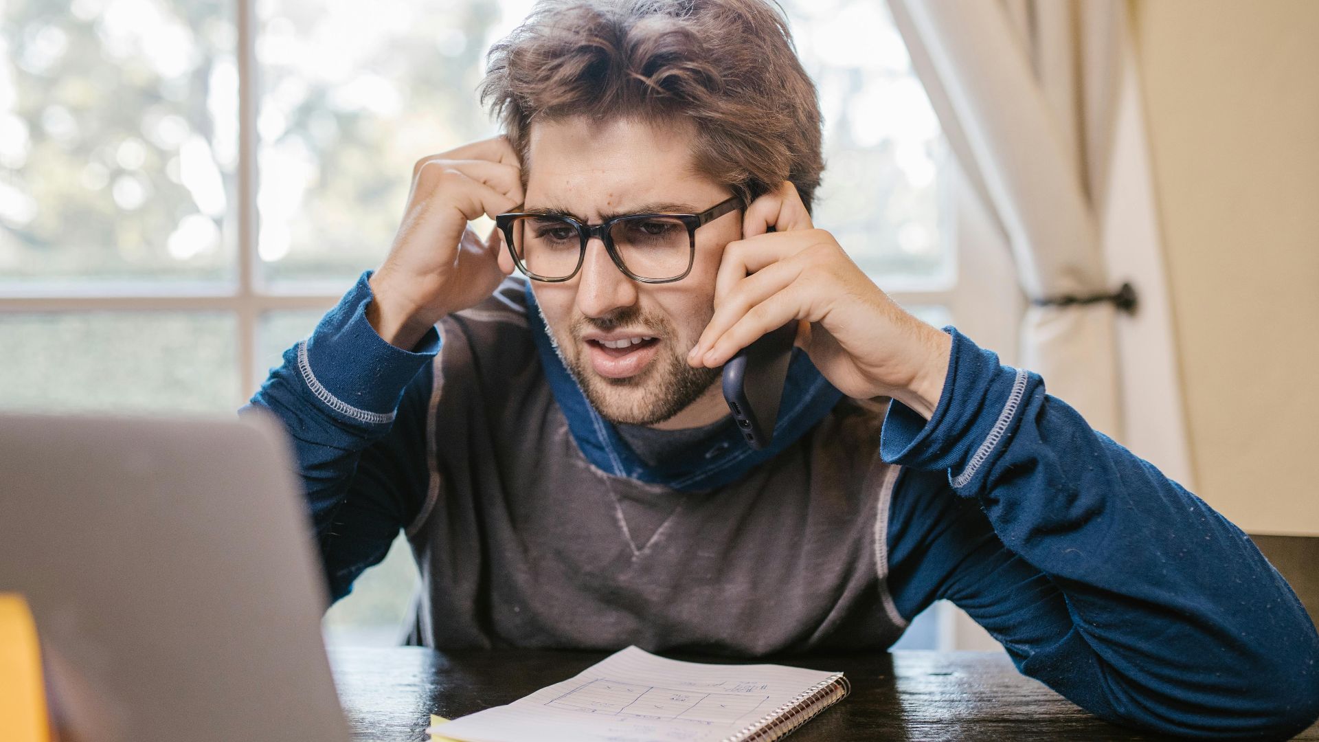 Man in glasses using laptop and smartphone at home, appearing confused.