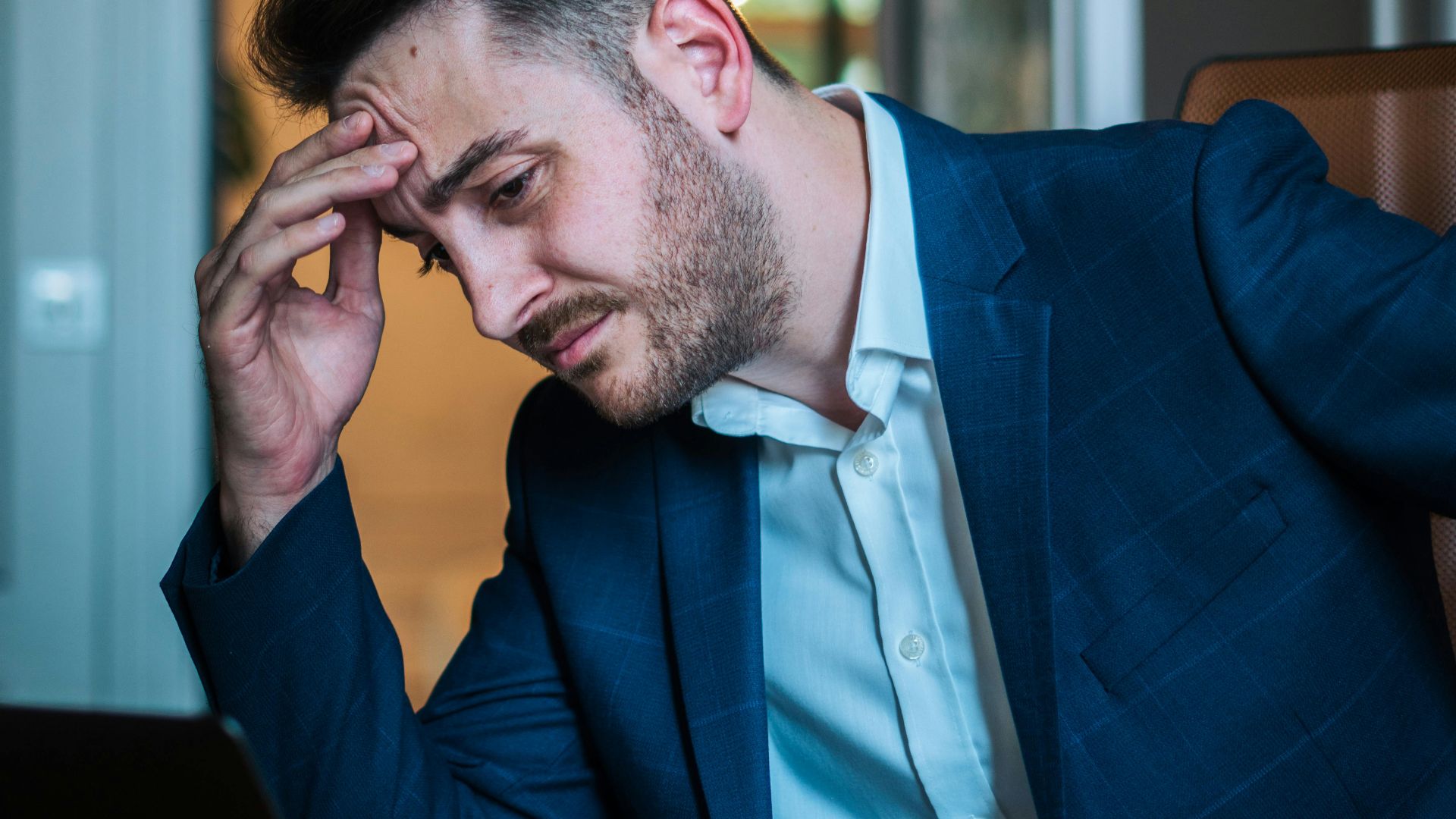A worried businessman in a blue suit working on his laptop in an office environment.
