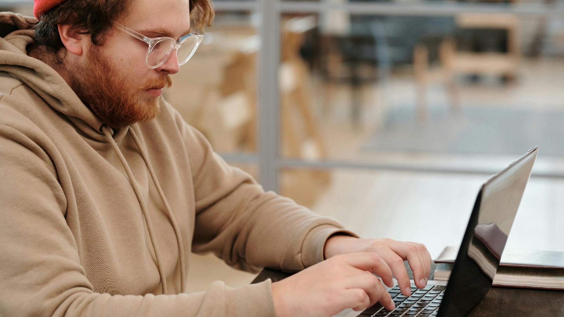 A bearded man wearing glasses, working intently on a laptop in a cafe setting.
