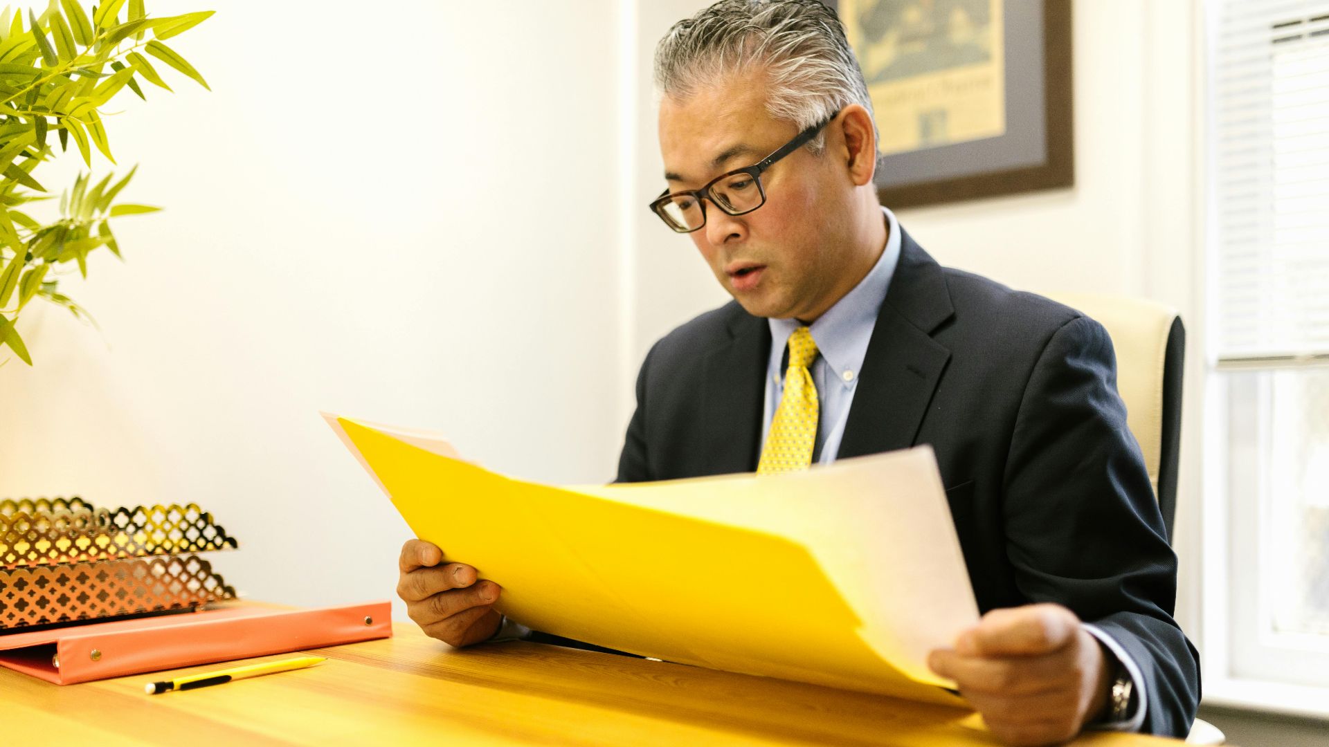 Asian businessman in corporate attire reading documents at office desk with a yellow folder.