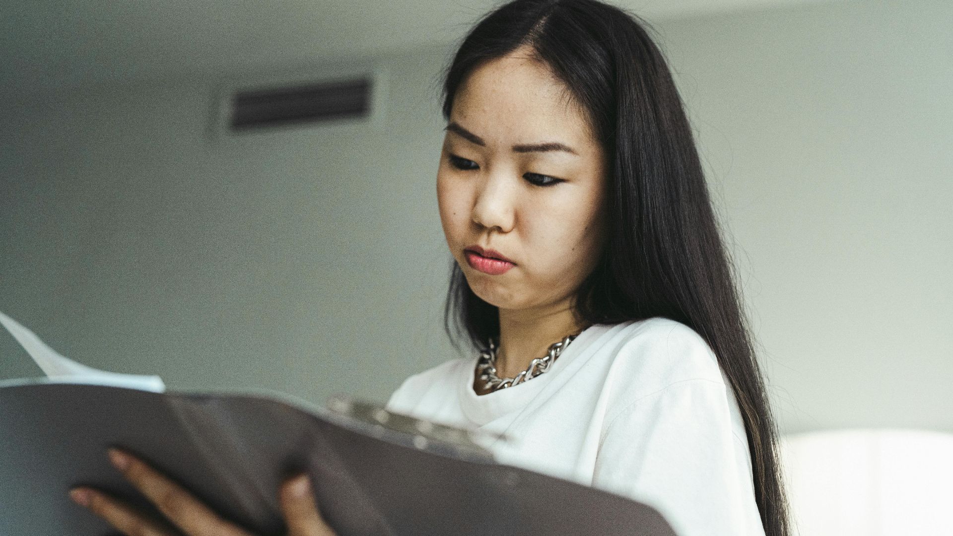 Asian woman in casual attire examining paperwork with concentration indoors.