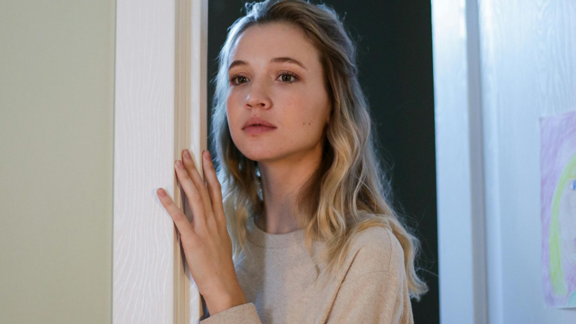 Young woman with blonde hair standing in a doorway, looking thoughtful indoors.