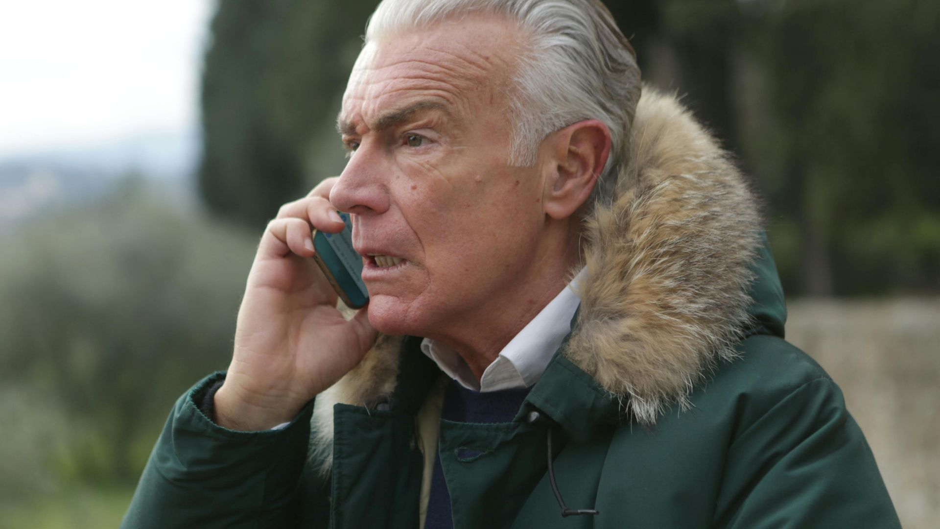 Elderly man in green coat talking on smartphone outdoors, showcasing lifestyle and technology.