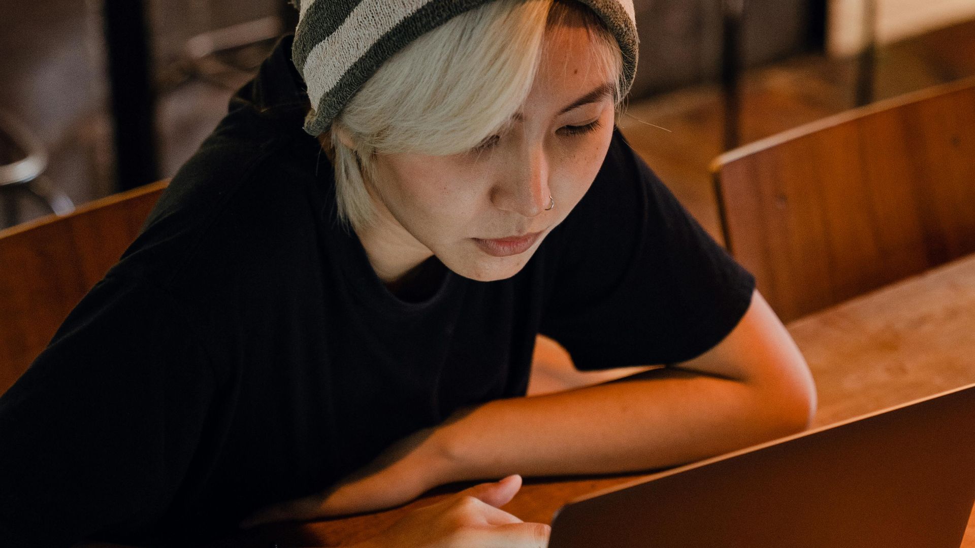 From above of young Asian lady with blond bangs in slouchy beanie and black T shirt sitting at table and browsing Internet sites on laptop in modern bar