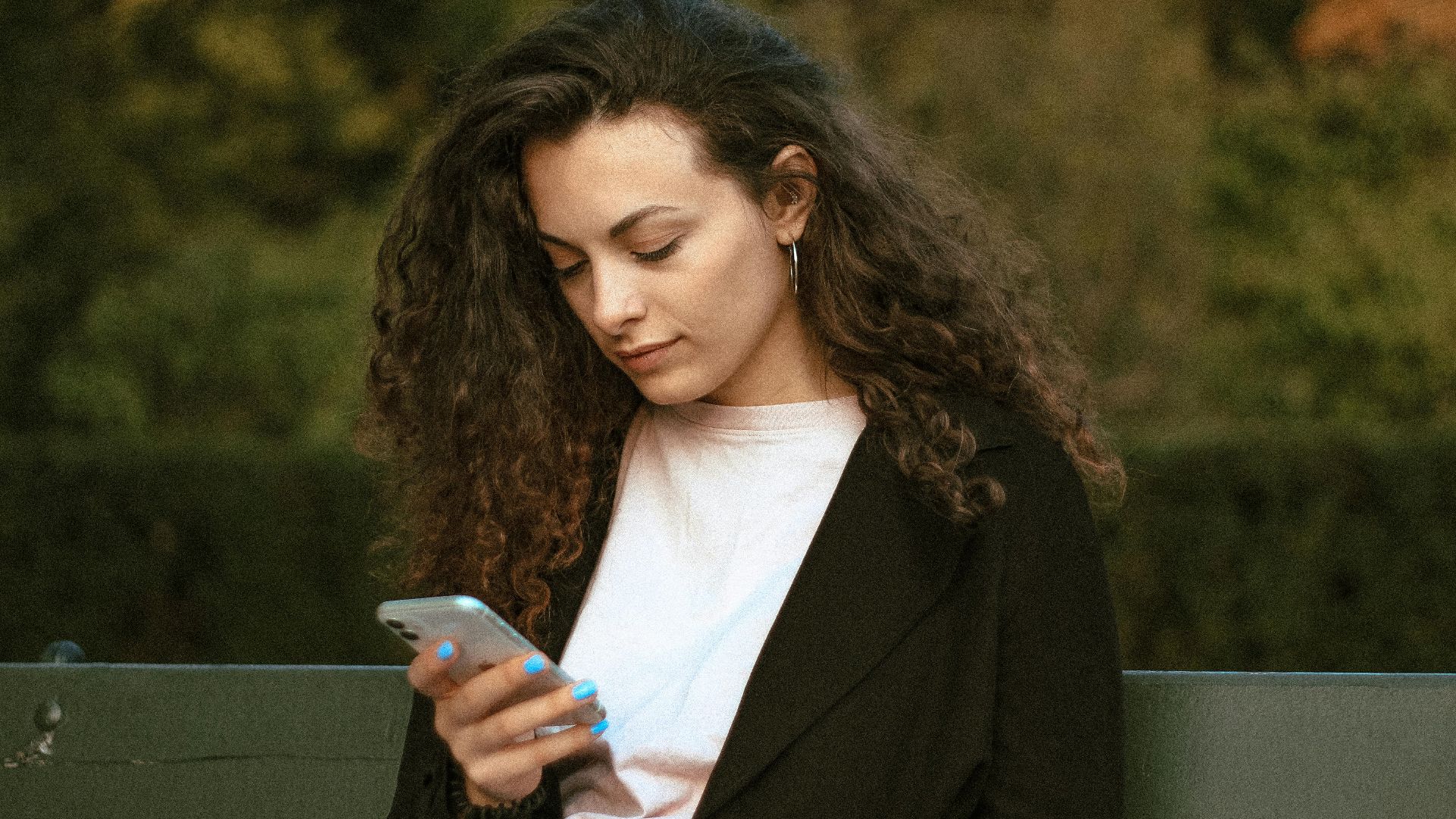 Woman with curly hair checking smartphone while sitting outdoors in Warsaw park.