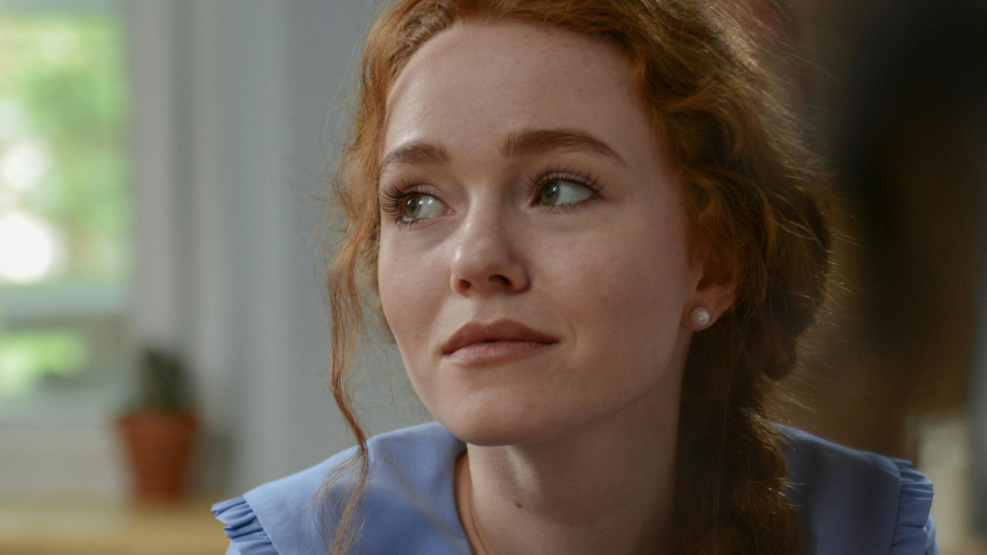 A red-haired woman in a blue blouse sits with crossed arms, looking pensively indoors.