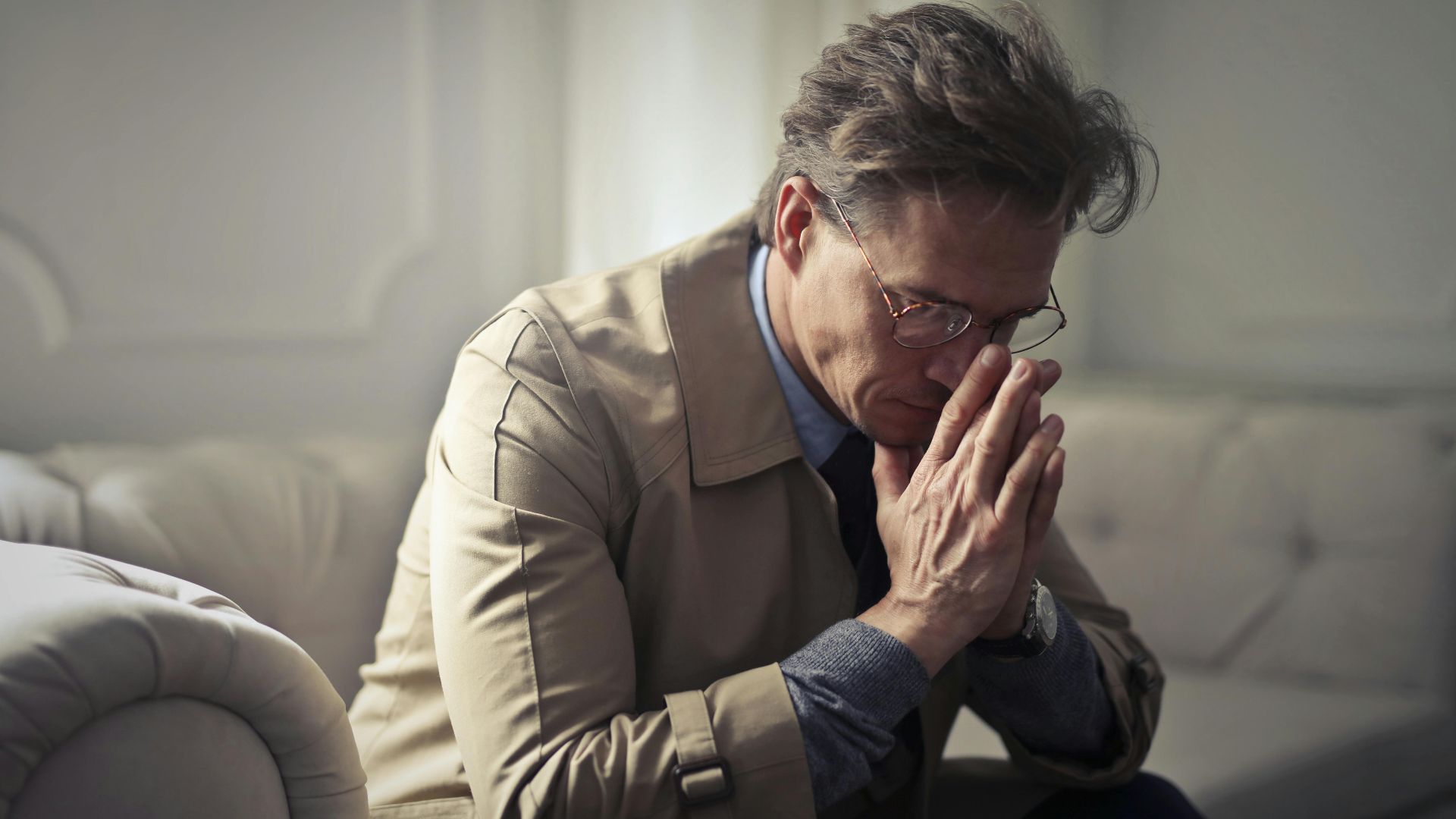 Side view of upset male entrepreneur in formal wear sitting on couch in modern living room and contemplating about financial problems while leaning on hands and looking down