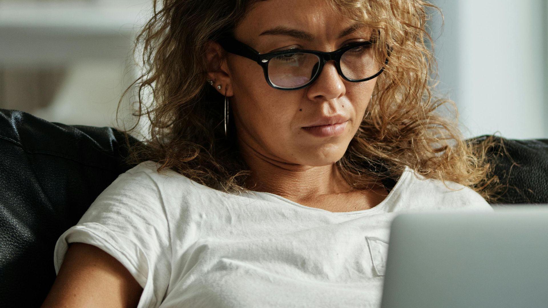 A woman with eyeglasses and curly hair working on a laptop while sitting on a sofa indoors.