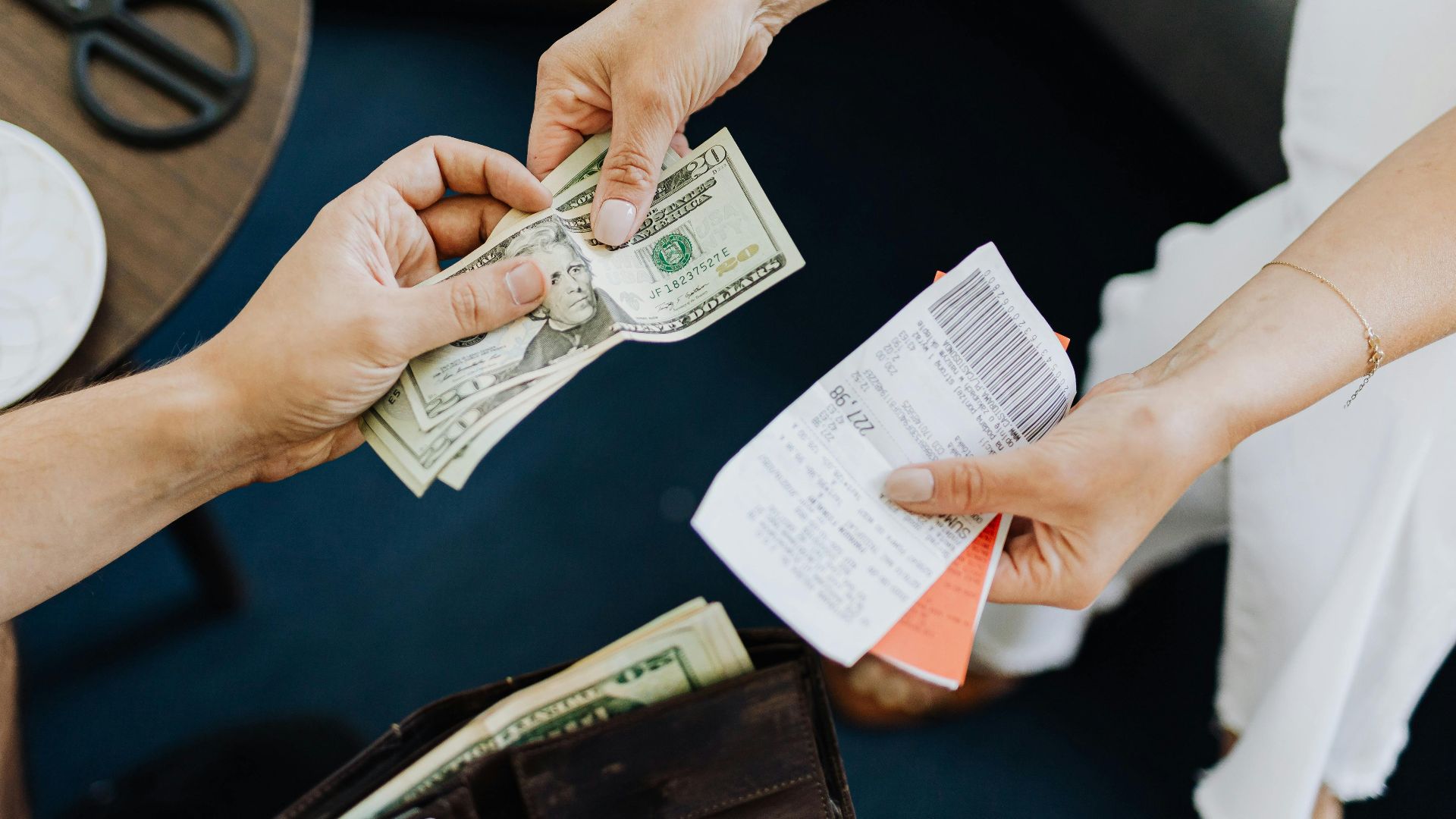 Close-up of a transaction with US dollar bills and receipts being exchanged over a table with coffee and scissors.