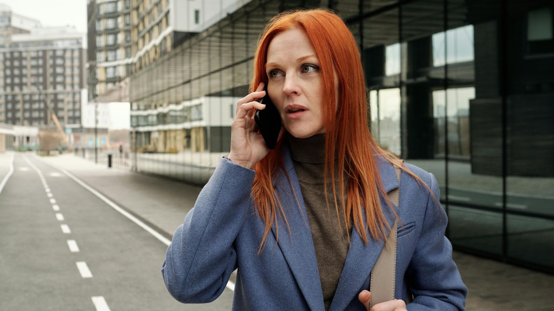 Red-haired woman in blue coat making a phone call on an empty city street.