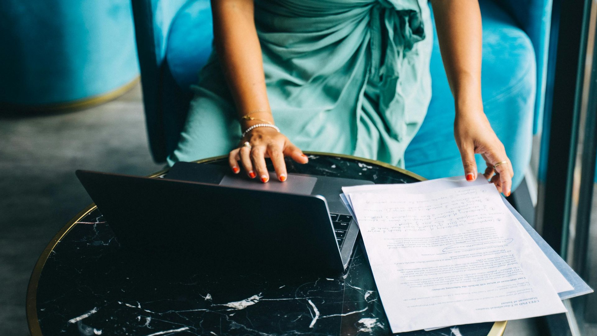 Adult woman in teal dress working on a laptop with documents, indoors.