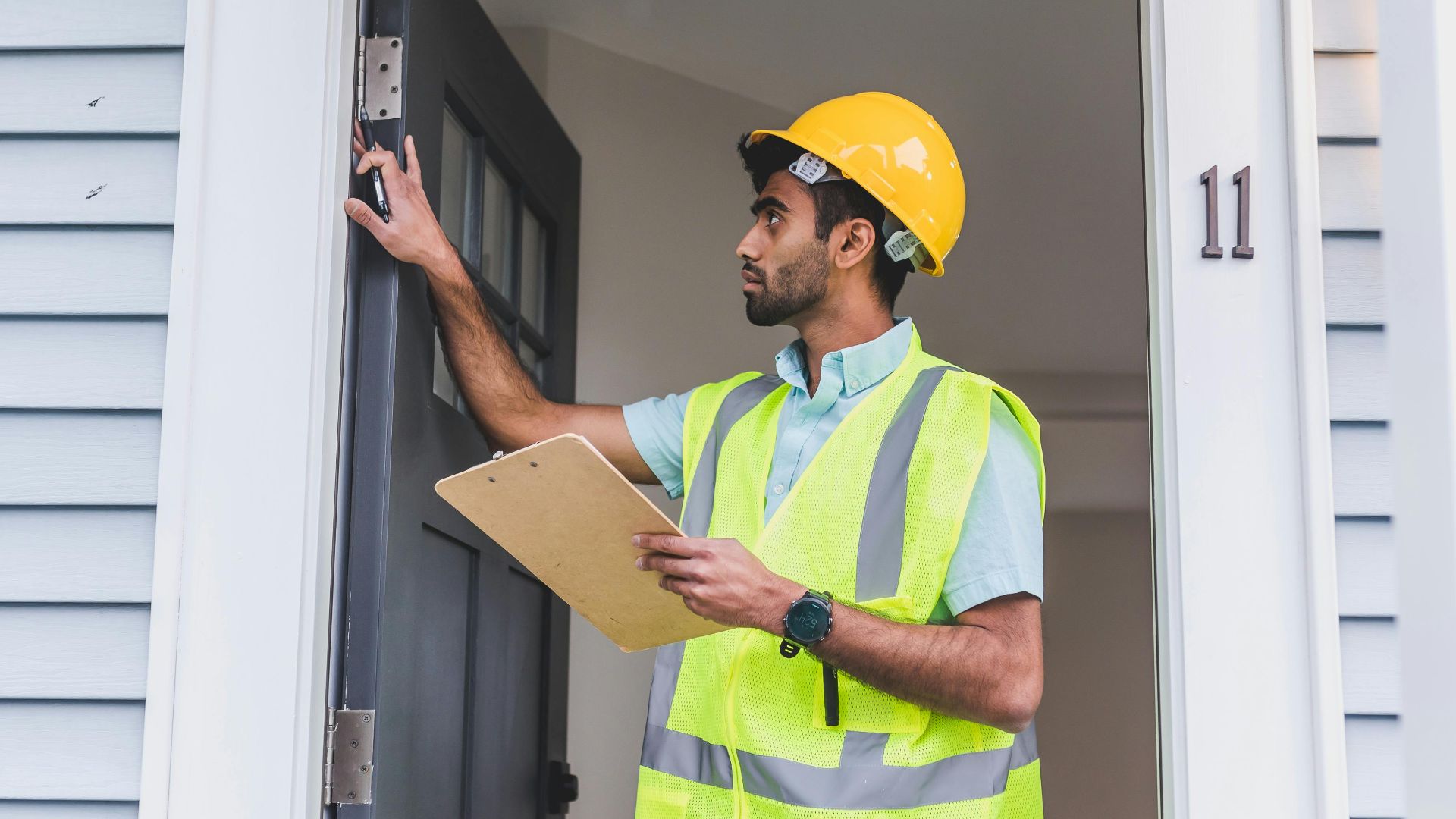 A home inspector in safety vest and hard hat checks doorframe alignment with clipboard outdoors.