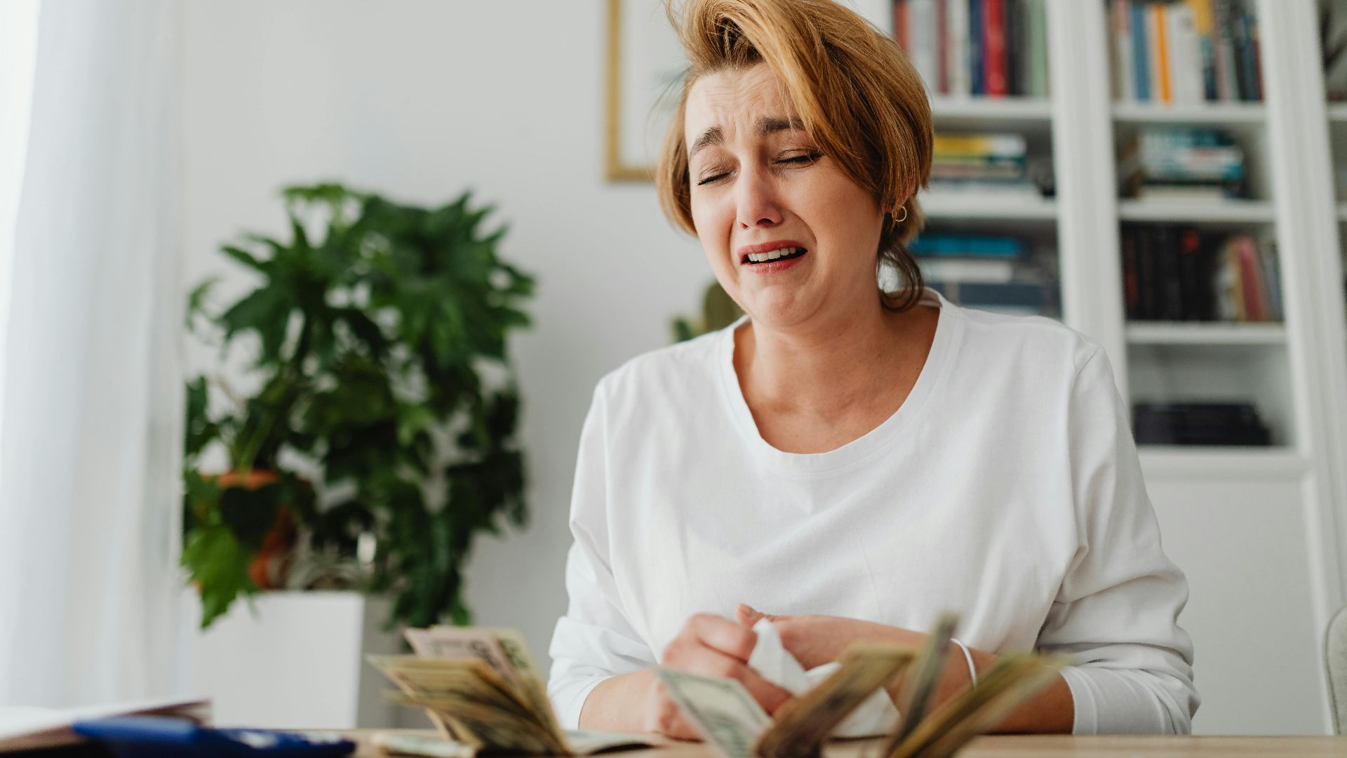 A distressed woman counts cash at a desk with a pained expression, highlighting financial strain.