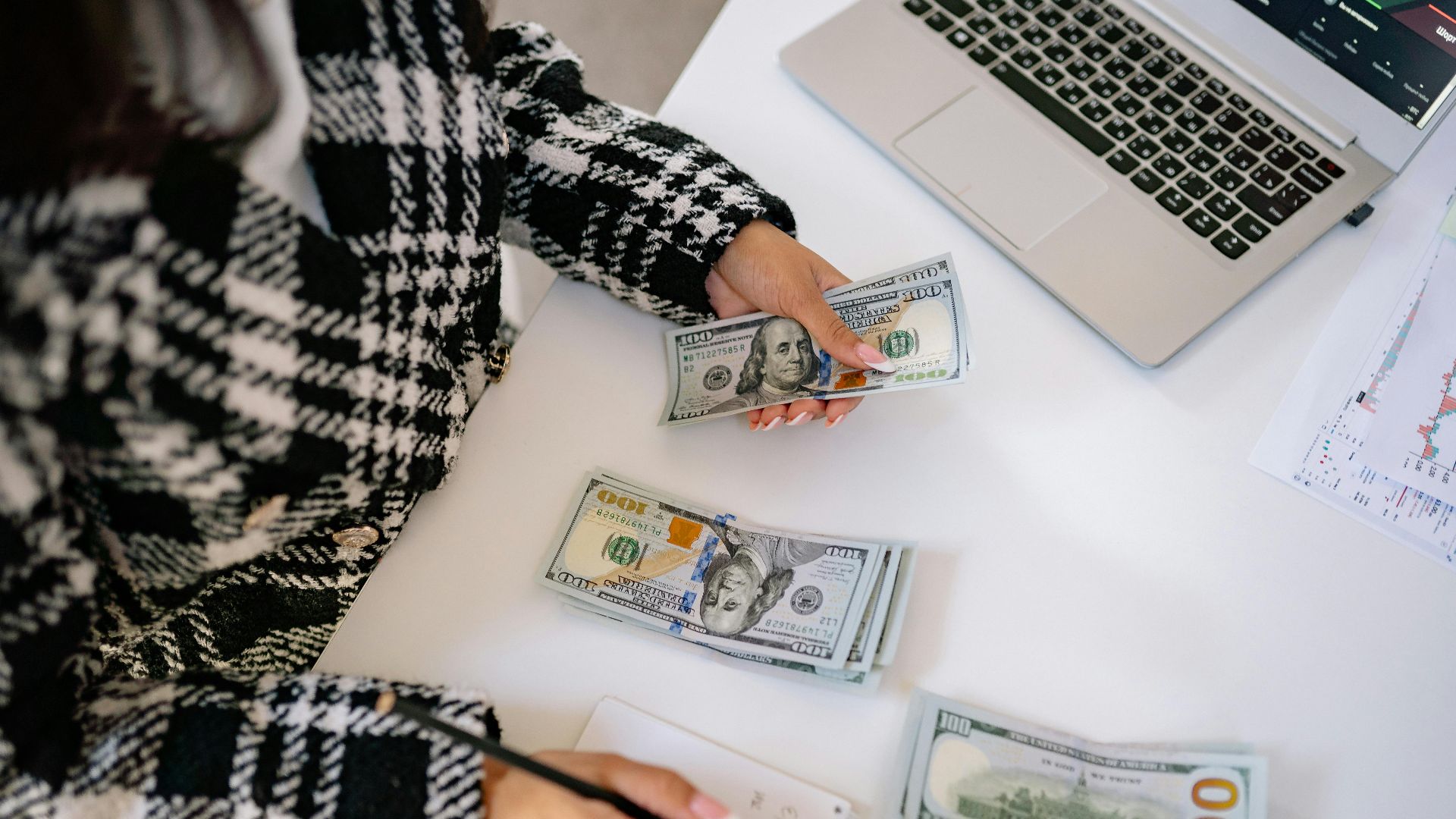 A woman in a plaid coat counts American dollars at a desk with a notebook and laptop.