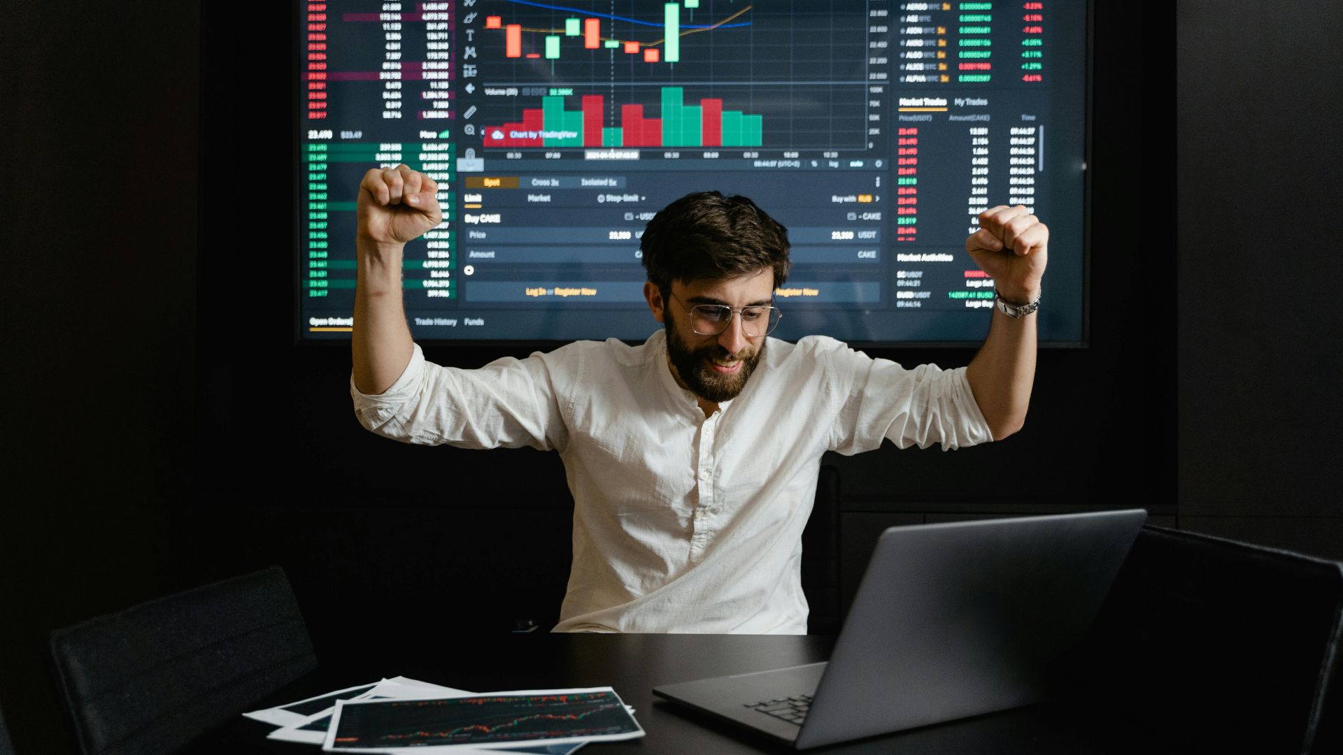 Businessman celebrates stock market success with hands raised in excitement at a trading desk.