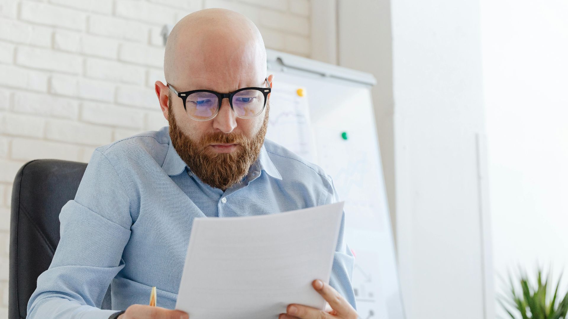 A businessman with a beard and eyeglasses reviewing documents in an office setting.