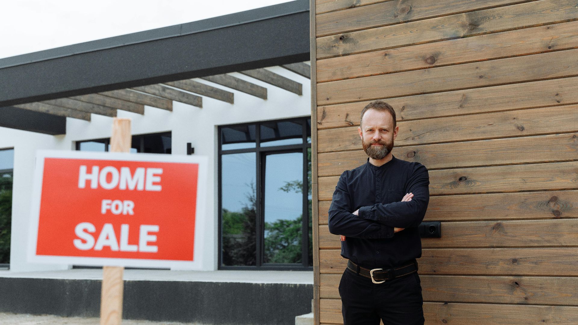 Real estate agent standing by a modern home with a 'Home for Sale' sign.
