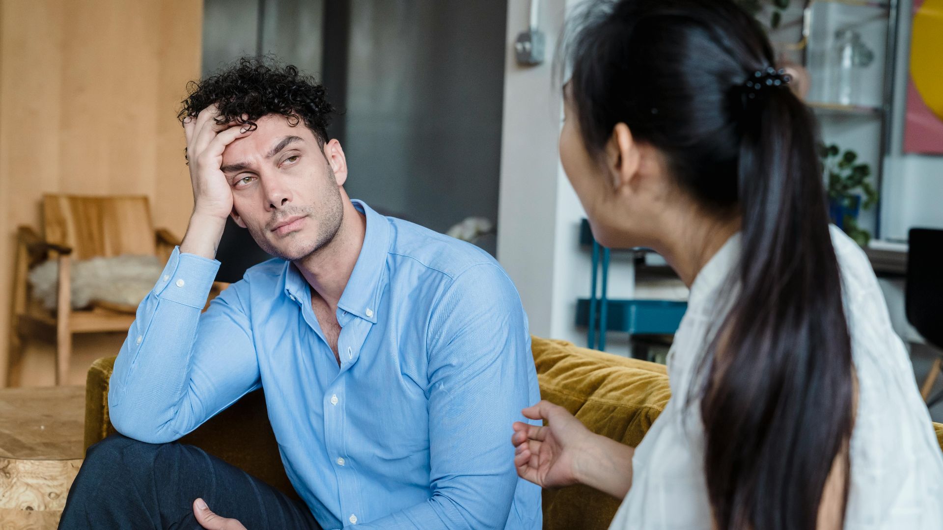 A tired man in a blue shirt listens while a woman talks indoors. Modern living room.