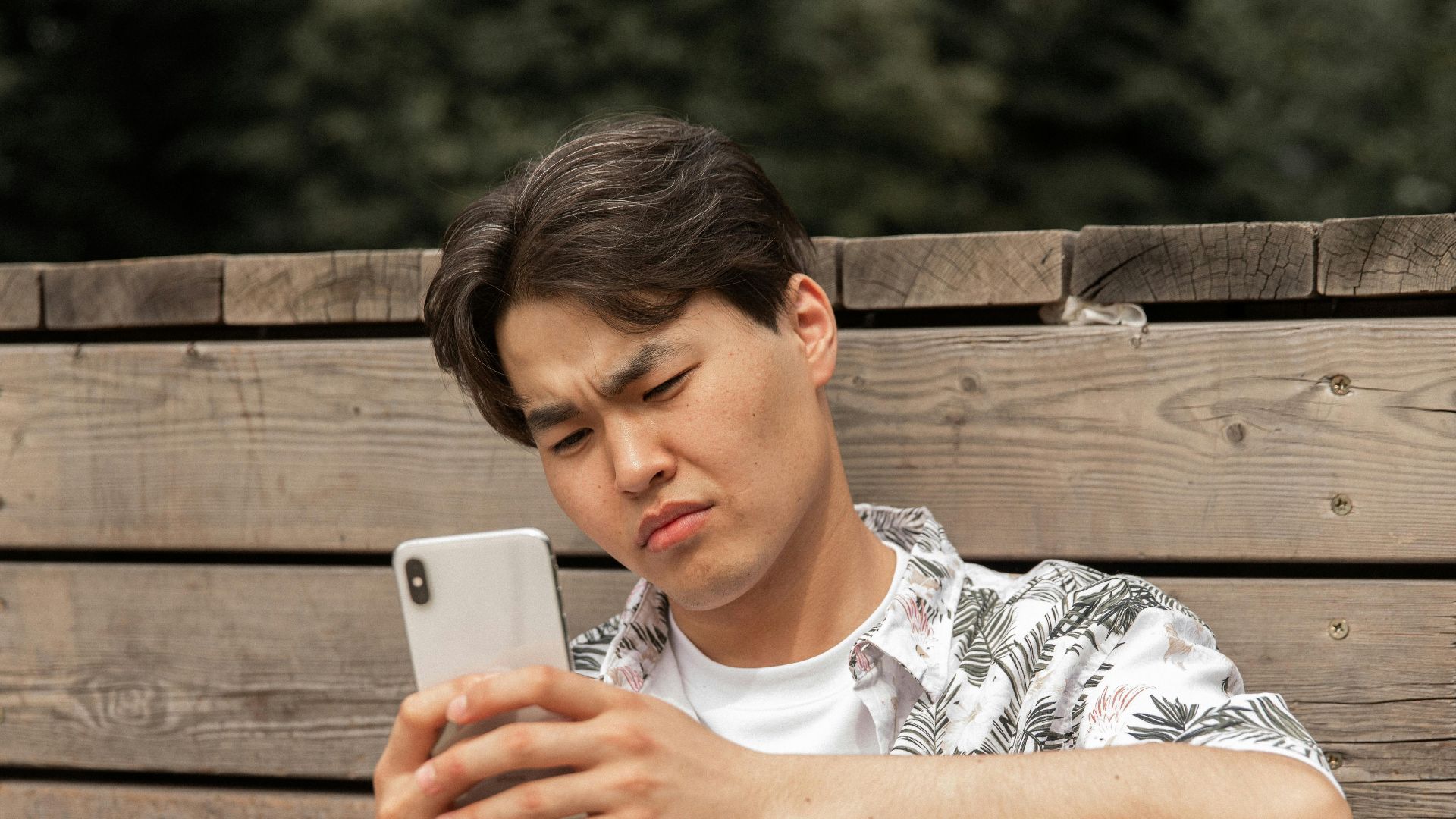 Young ethnic male in stylish clothes leaning on wooden barrier while messaging on mobile phone in park