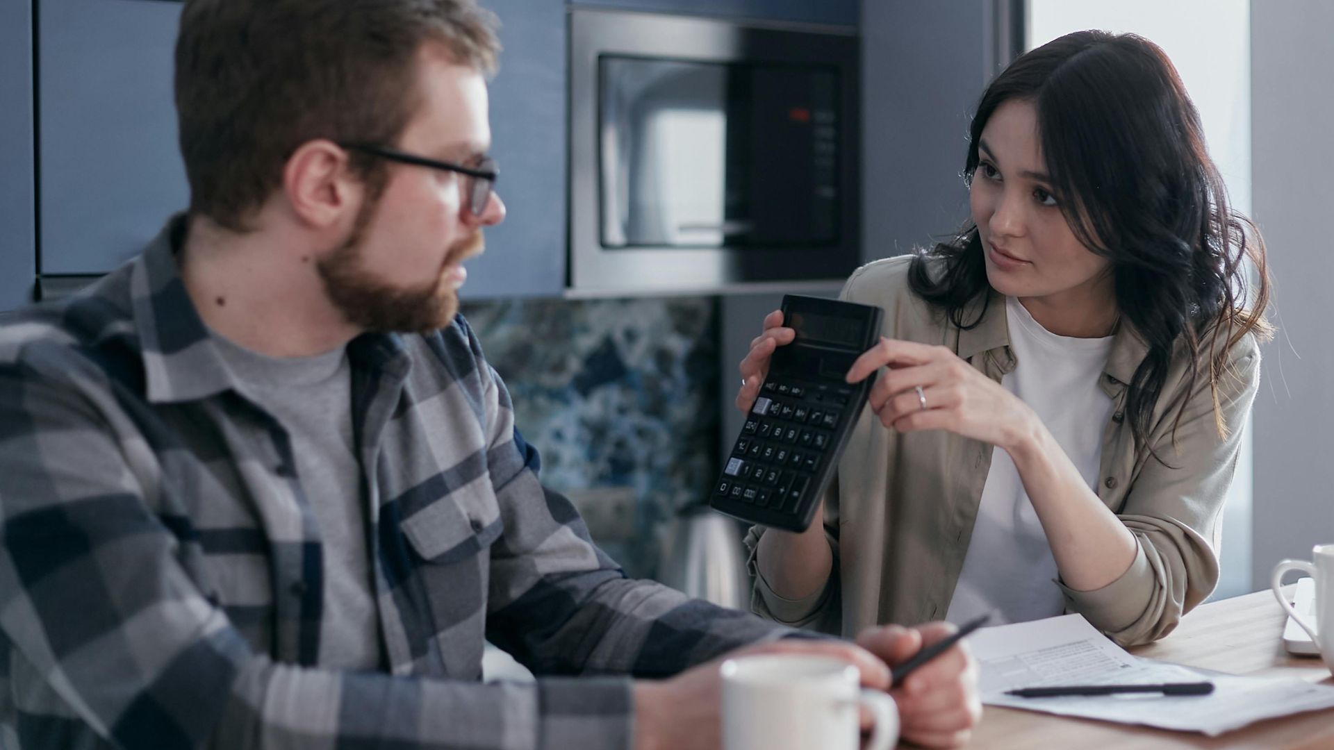 A couple discussing finances with a calculator and papers at a wooden table, looking concerned.