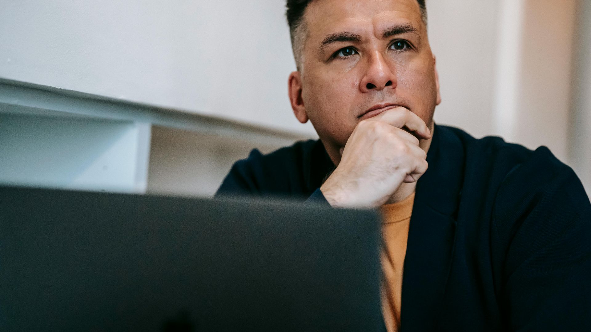Focused man in home office pondering work on a laptop, expressing deep concentration and thought.
