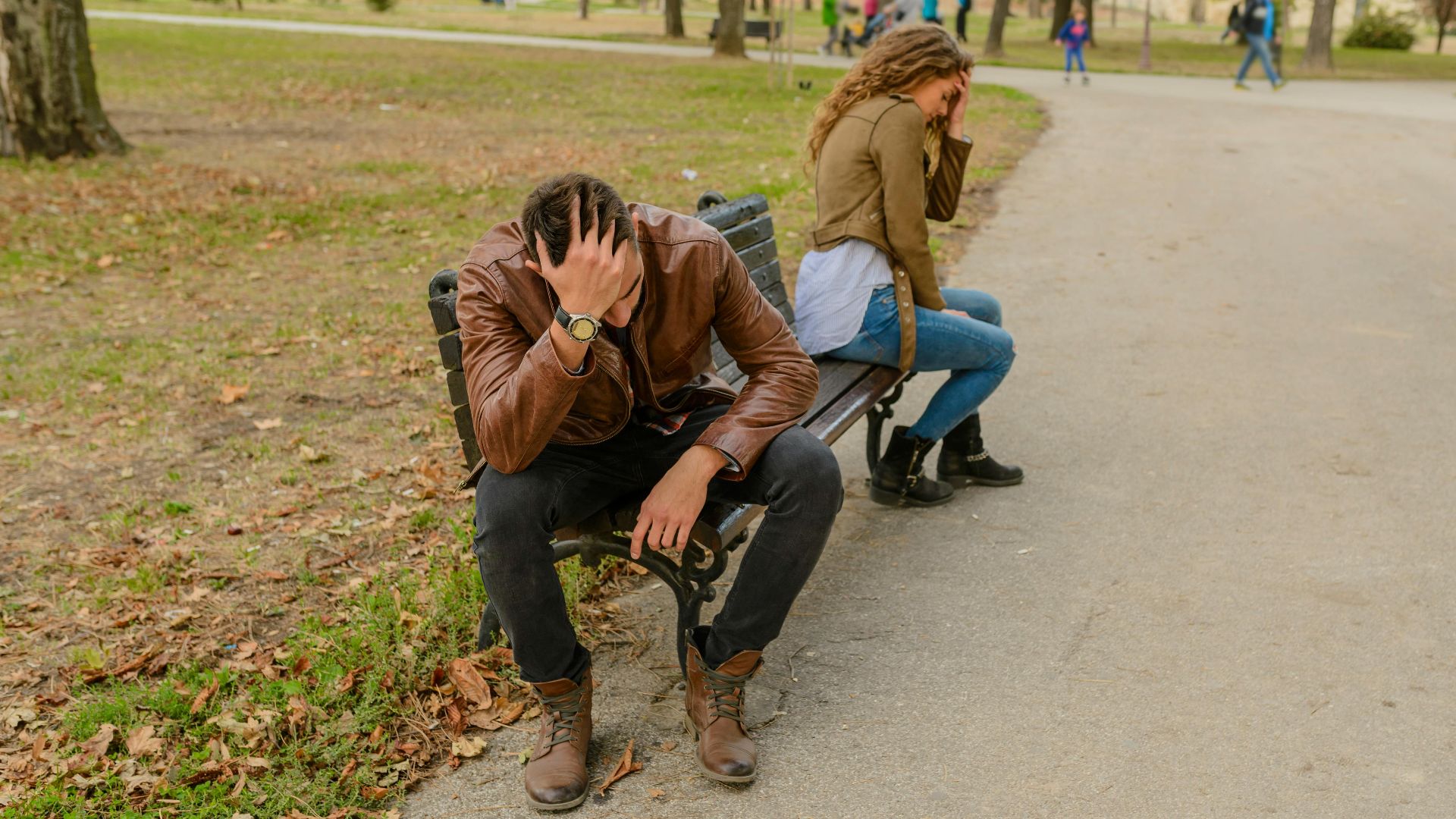 Distraught couple sitting apart on a park bench, facing away from each other, in a public park setting.