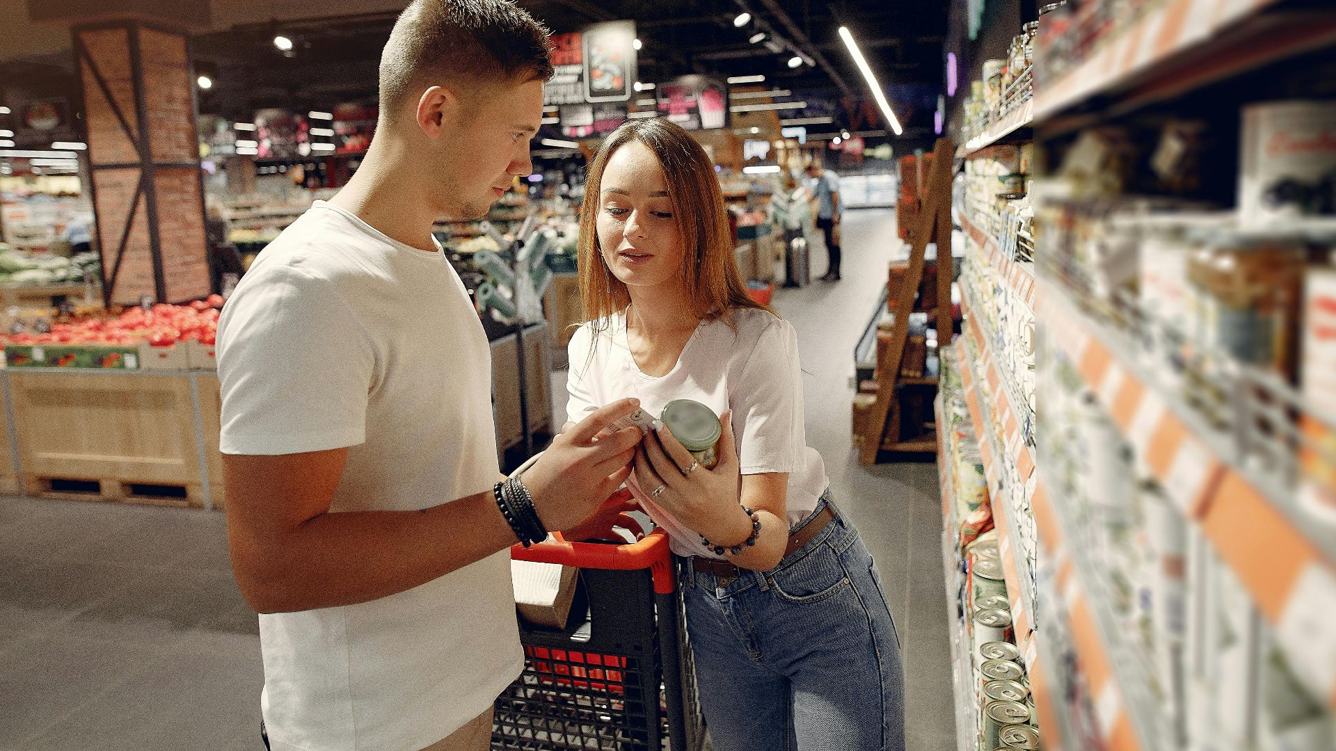 From above of young woman in casual wear showing canned food for man while standing with grocery trolley near shelves with products in modern supermarket