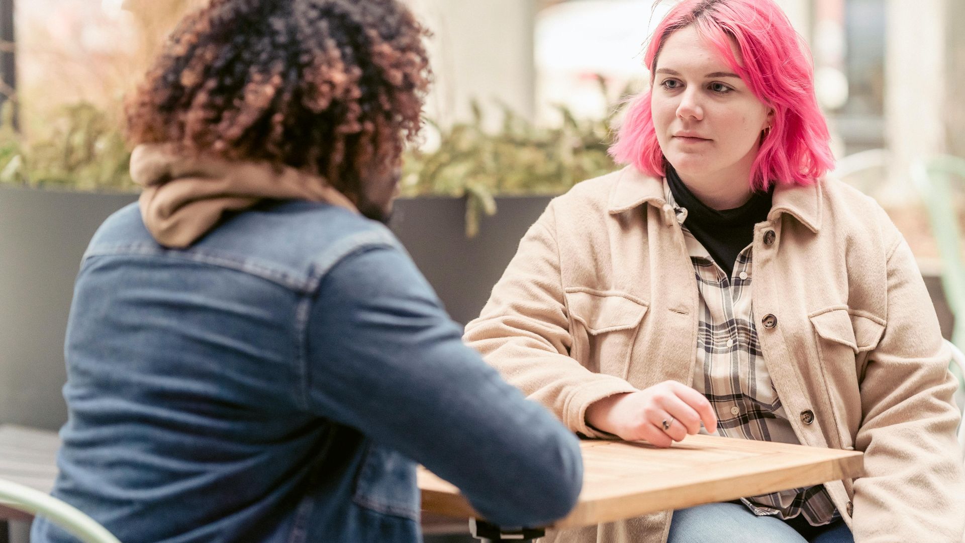 Young woman with pink hair listening to black man while sitting at table in weekend in outdoor cafeteria