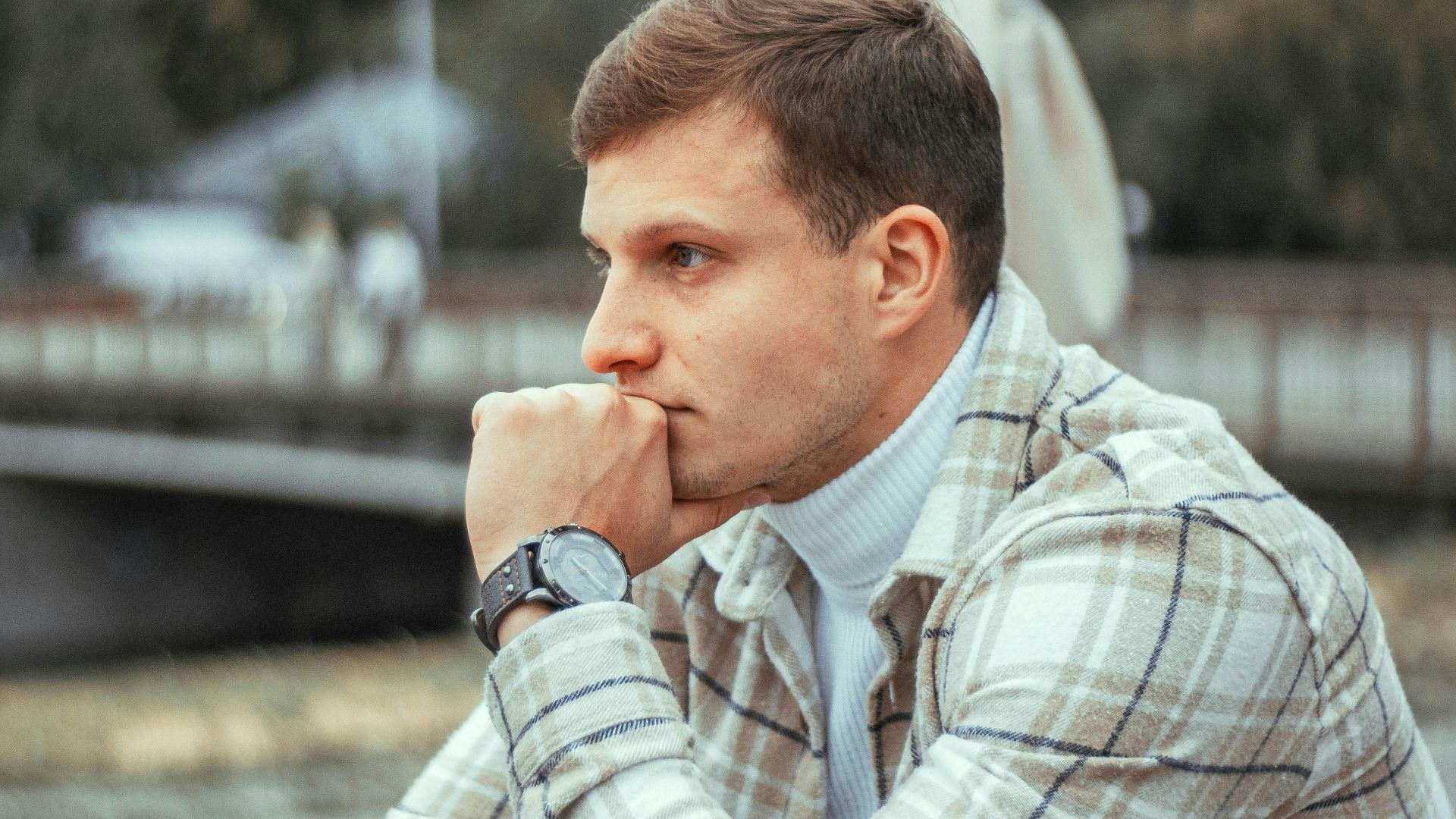 Close-up of a man in a plaid jacket, deep in thought at an outdoor cafe in a city.
