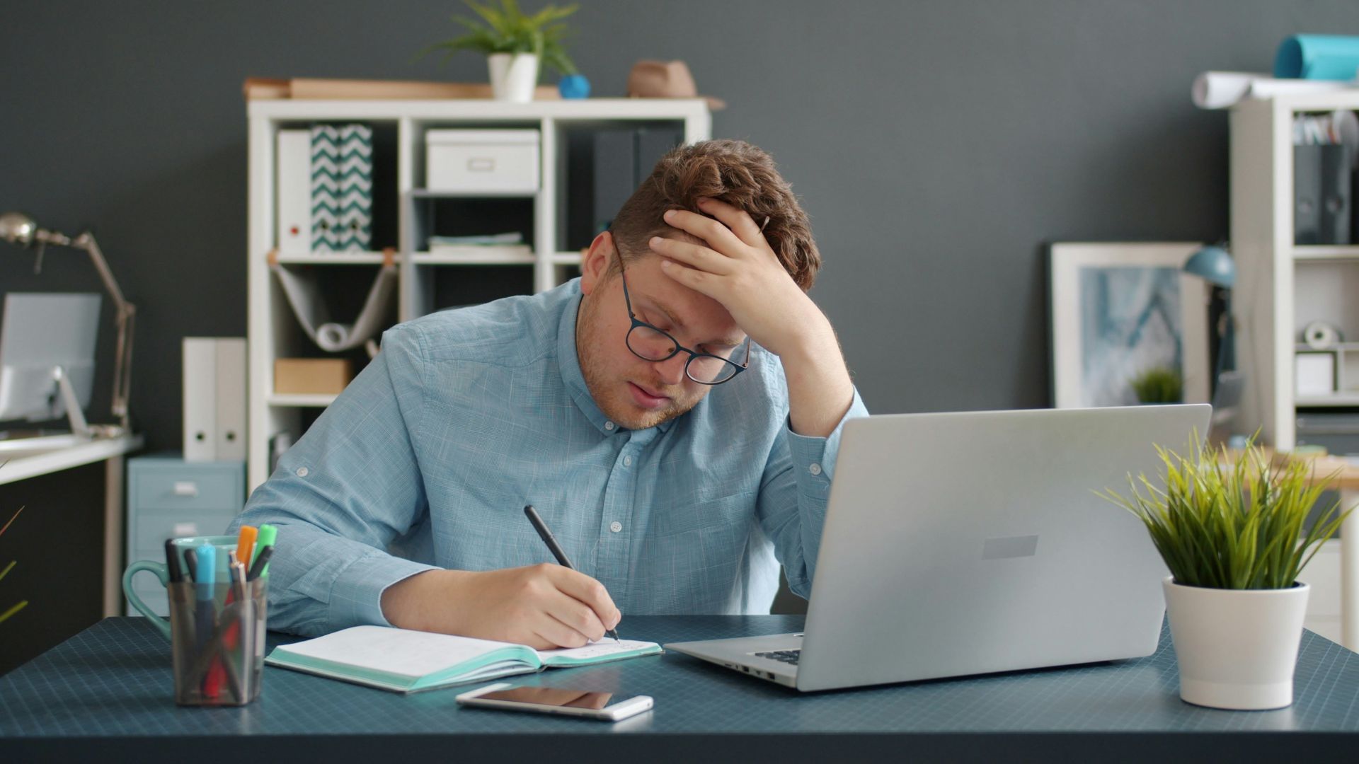 Man writing at desk with laptop, looking stressed.