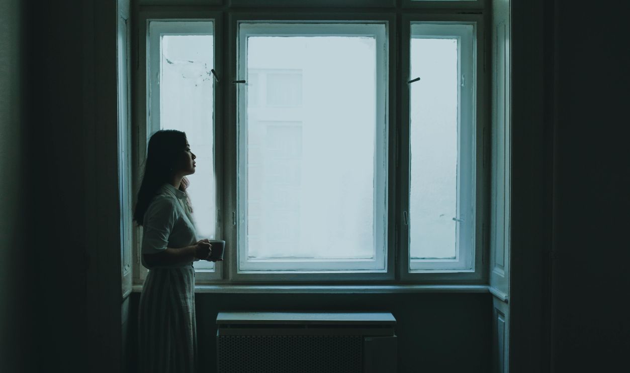A young woman with long hair stands by a window holding a cup, creating a serene and contemplative mood.