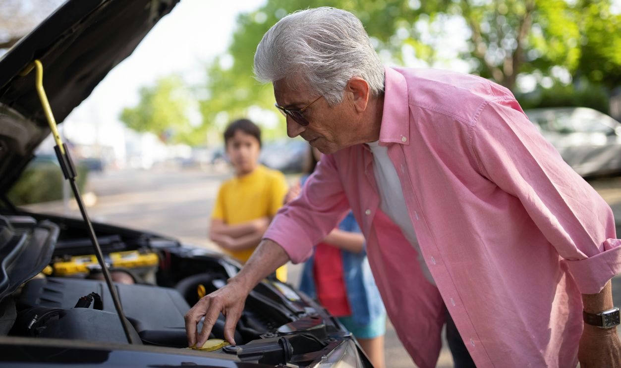 Elderly man inspecting car engine on a sunny day with children nearby.