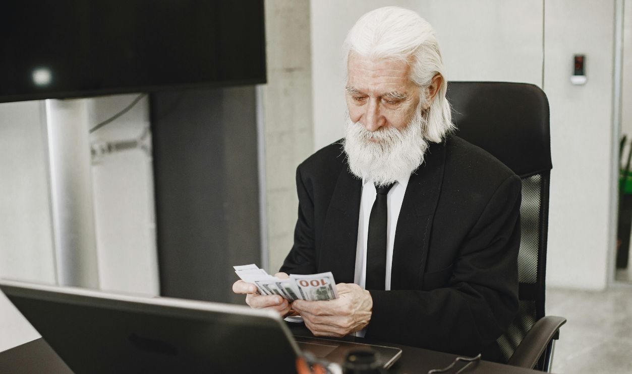 Senior businessman in suit counting money at office desk with a laptop.