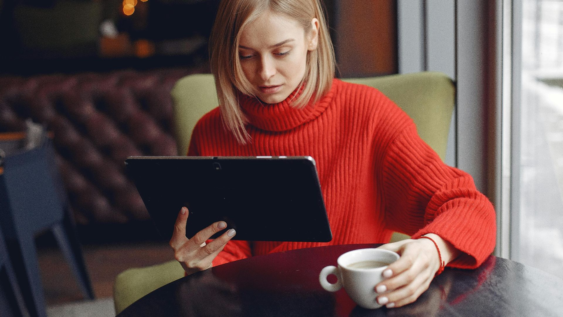 Woman in a red sweater sitting and using a tablet with a cup of coffee in a cozy cafe