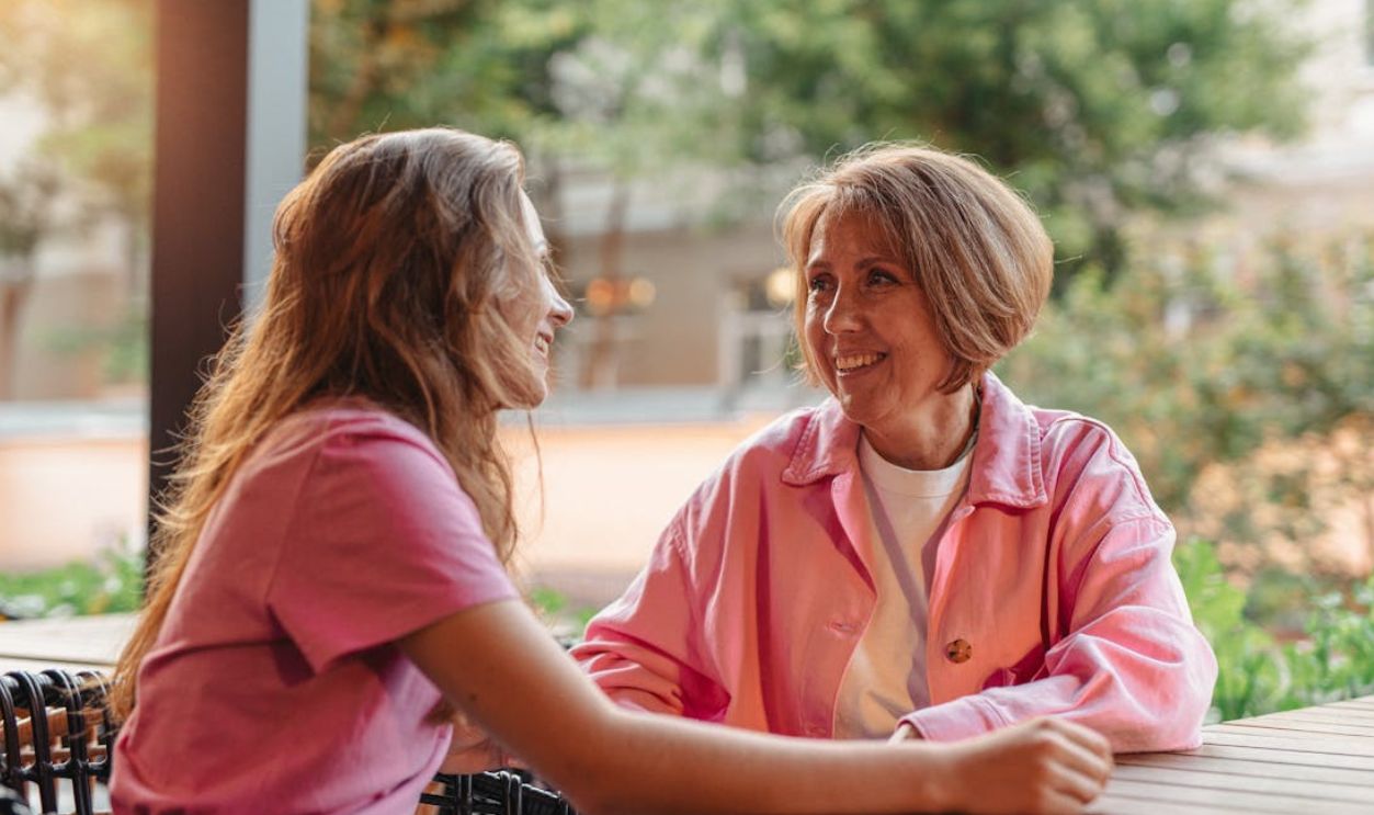 Two women smiling and chatting at a wooden table outdoors, depicting friendship and connection