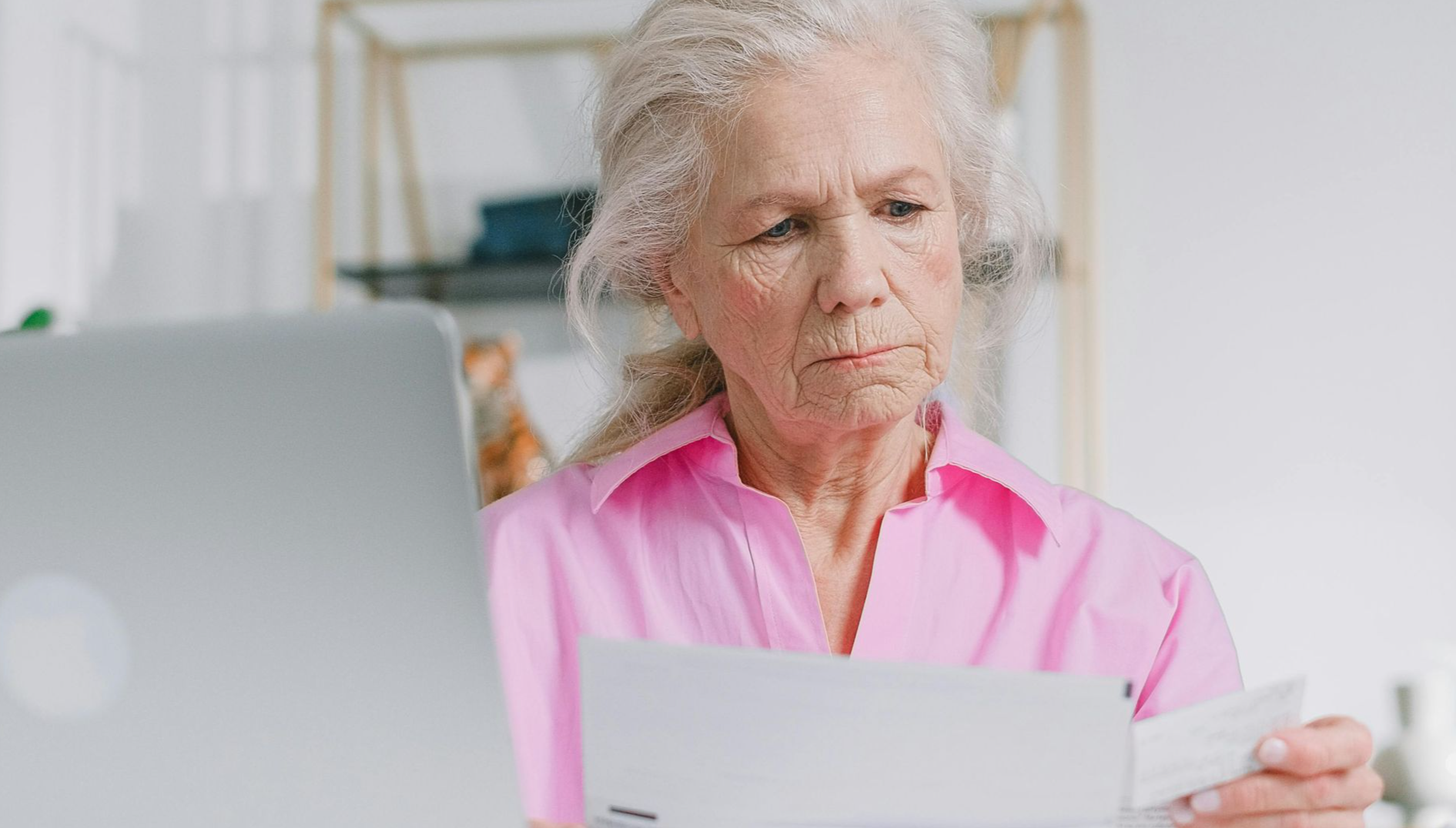 Elderly woman in pink blouse reviewing documents at home office desk.