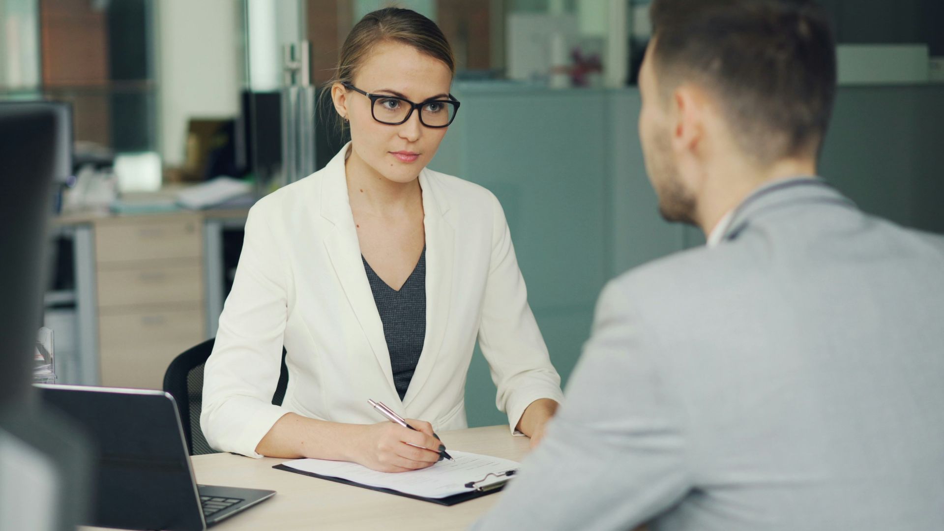 Woman in glasses interviews man at office desk.