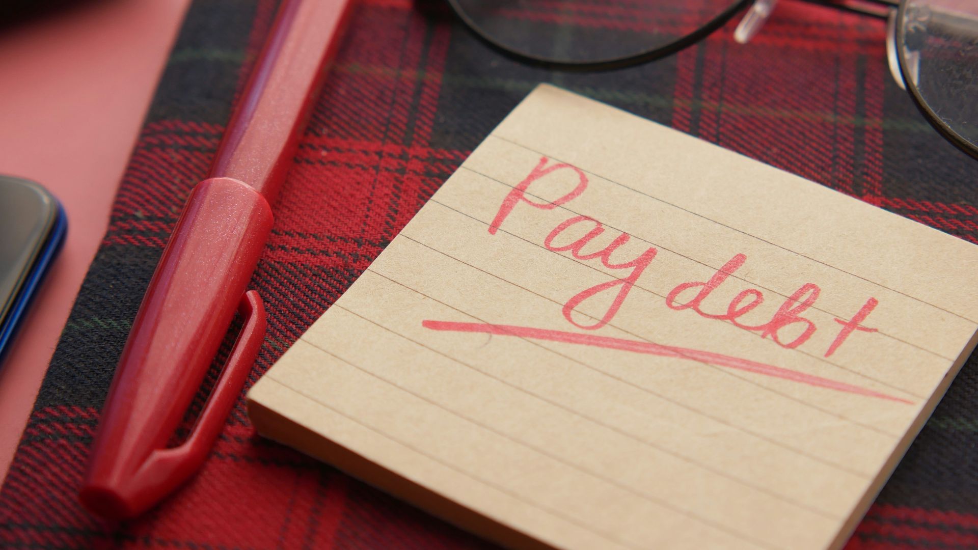 a note that says pay debt next to a pen and glasses