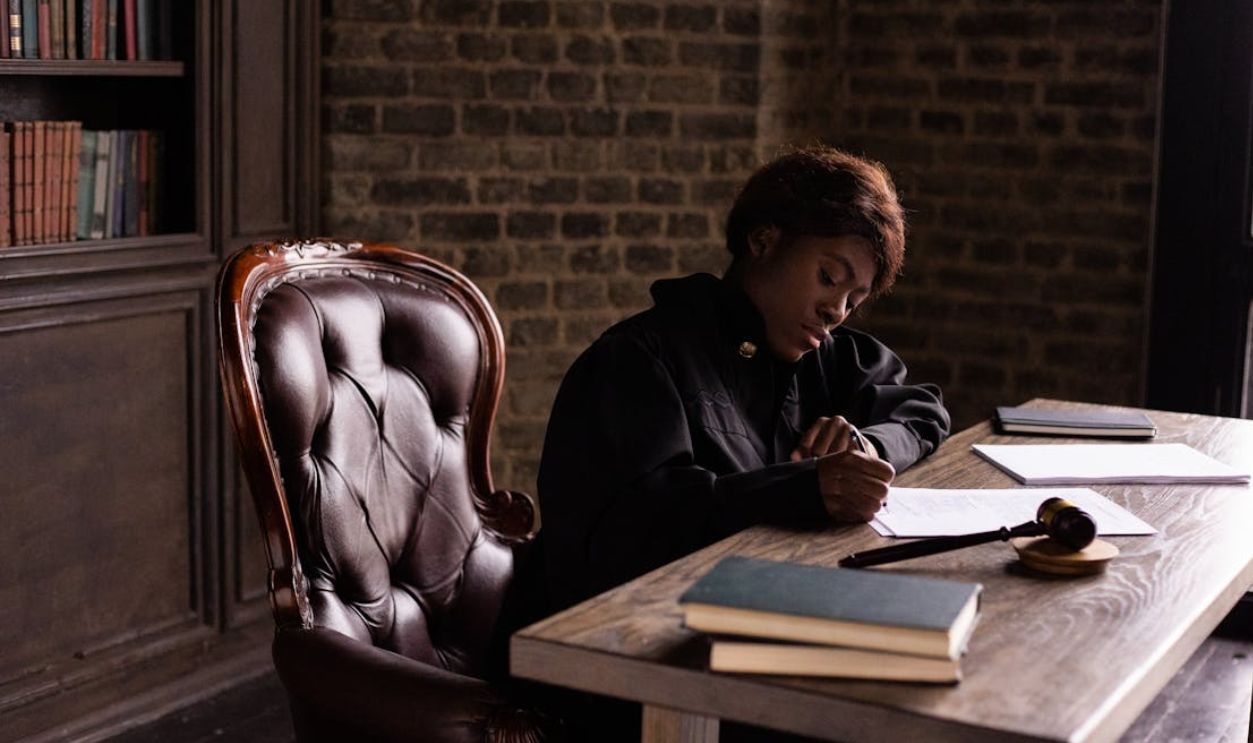 Black female judge in judicial robe writing at desk in an office setting, focusing on legal work